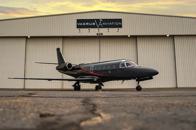 A dark gray jet with orange stripes parked in front of a hangar with the Vaerus Aviation logo.