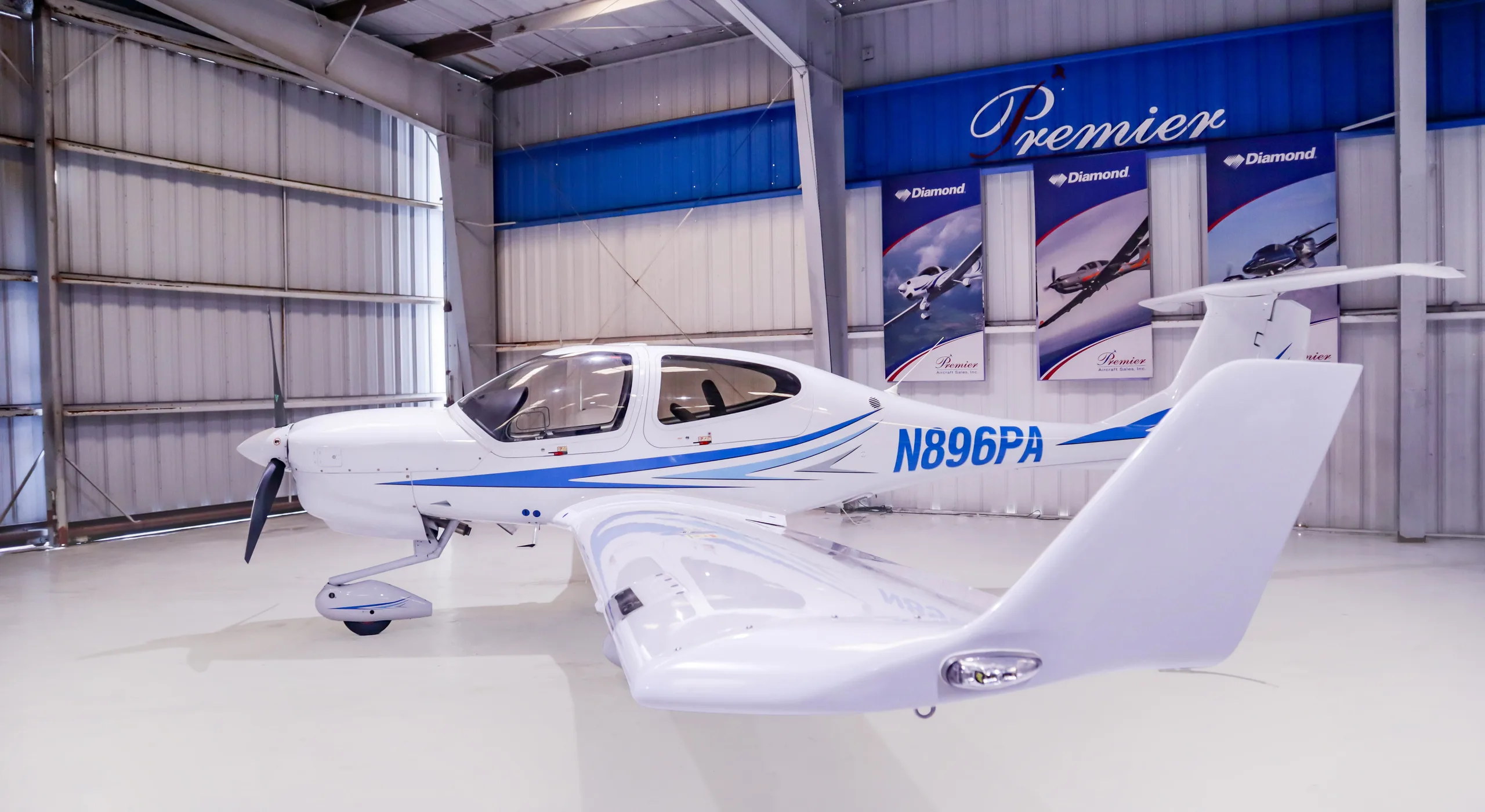 A rear-right view of a white Diamond DA40 aircraft with blue stripes, registration N896PA, parked inside a hangar.