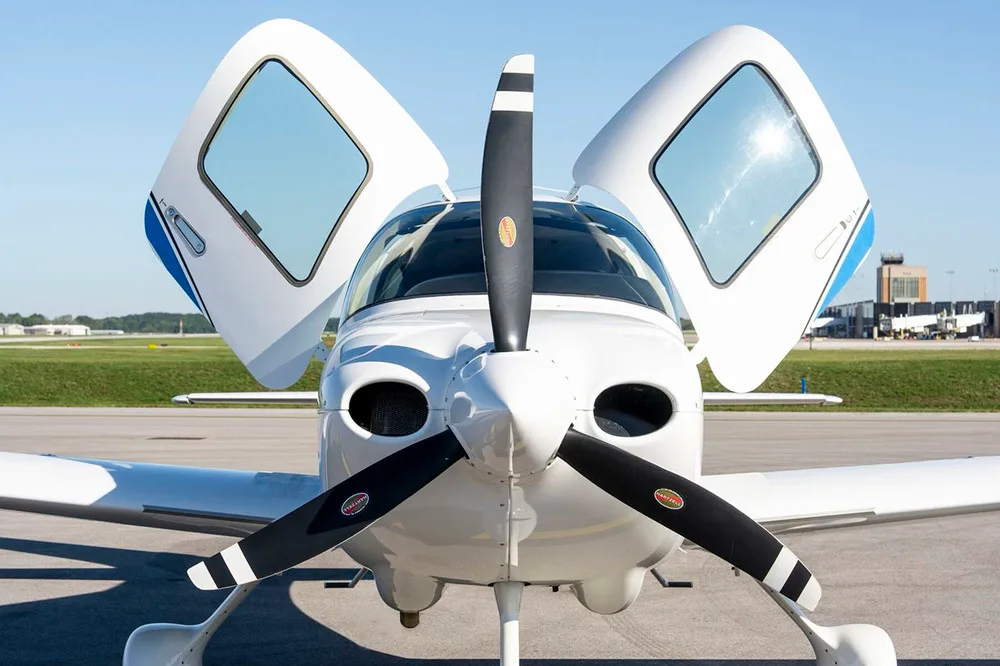 A front close-up view of the white Cirrus SR22 aircraft with its gull-wing doors open, showing the propeller.