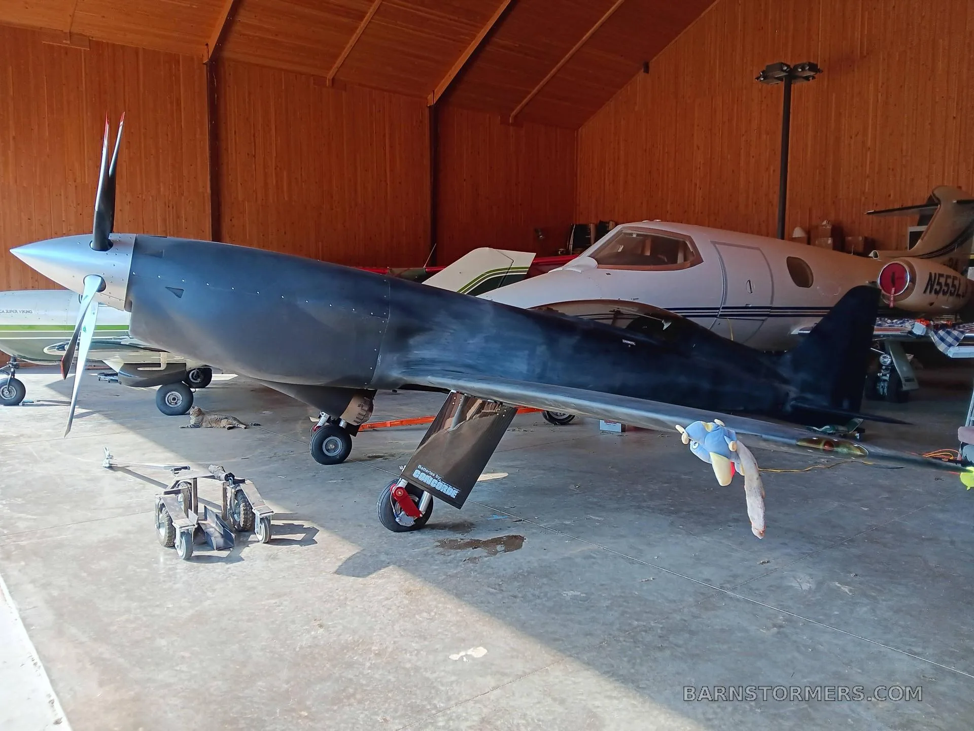 A dark, single-engine aircraft with a silver spinner and propeller, parked inside a wooden hangar next to another white aircraft.