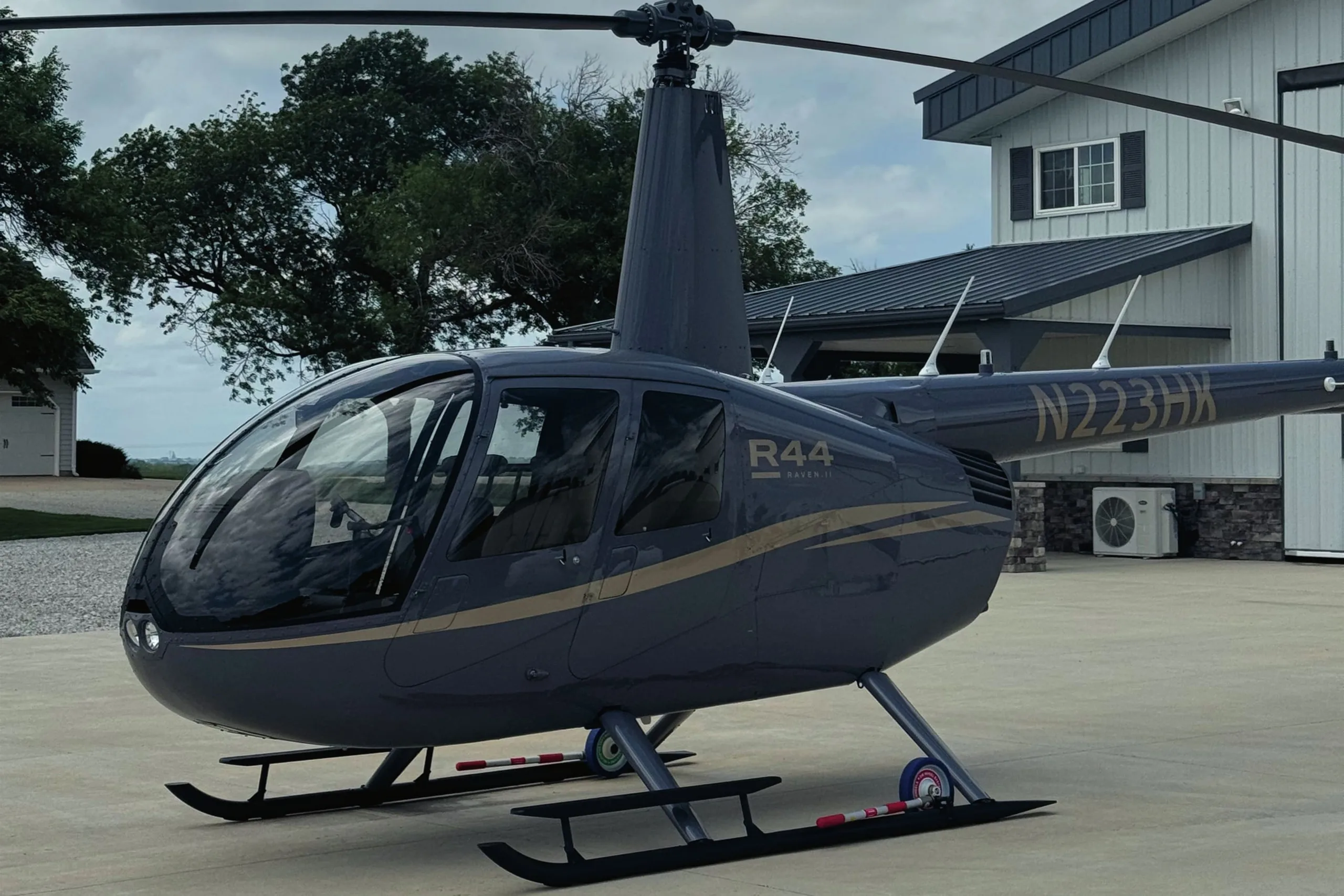A dark grey Robinson R44 Raven II helicopter with gold stripes sits on a concrete pad in front of a modern building and trees.