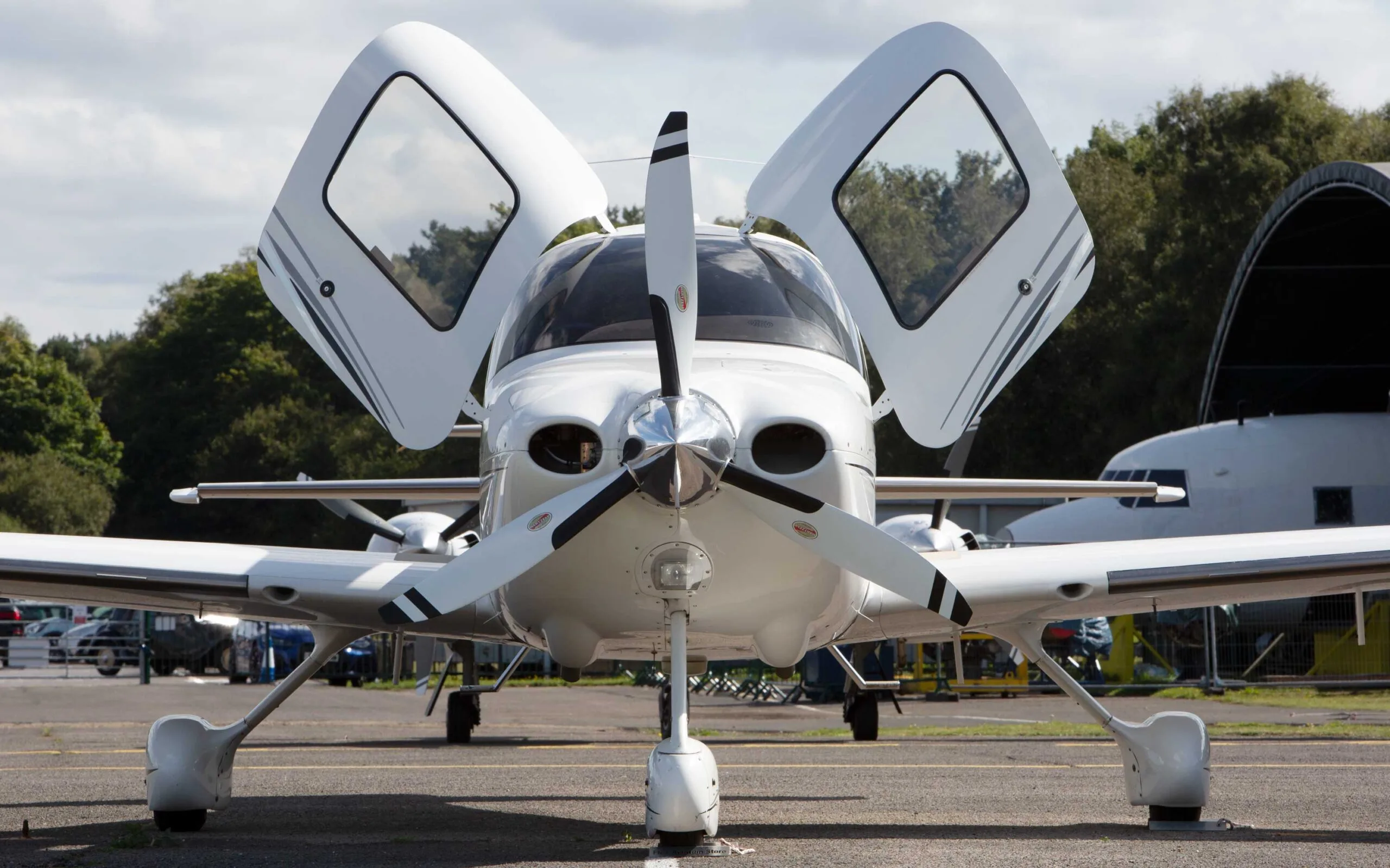 A white Cirrus SR22 TSi aircraft with black and grey stripes, registration G-YEBO, seen from the front with its gull-wing doors open on an airfield.