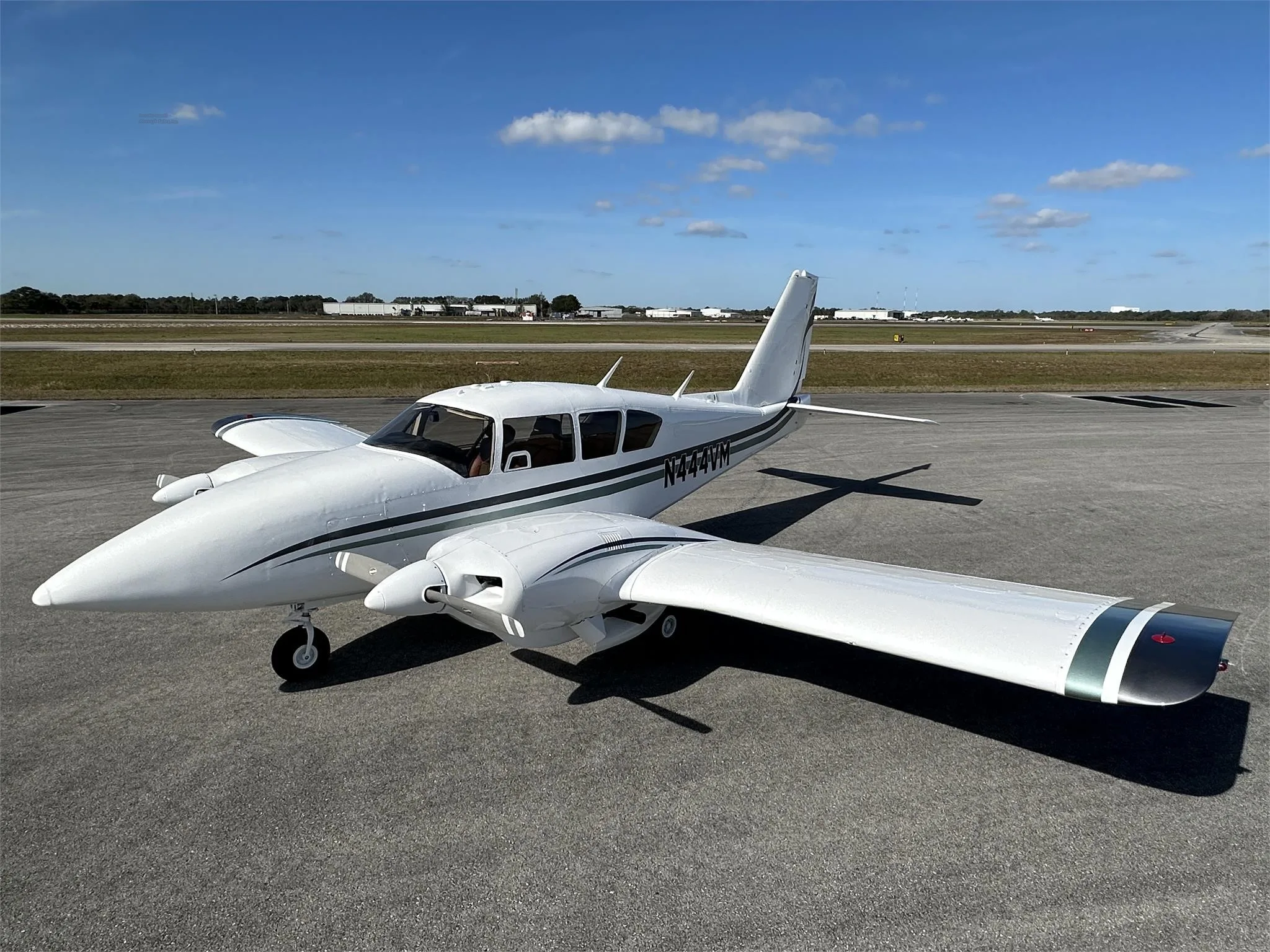 A white Piper aircraft with green and black stripes, registration N444VM, parked on an airfield under a blue sky.