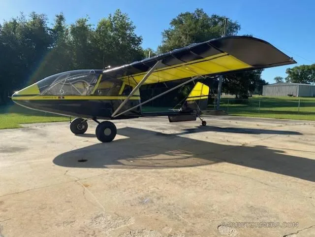A black and yellow ultralight aircraft with a high wing, parked on a concrete pad outdoors.