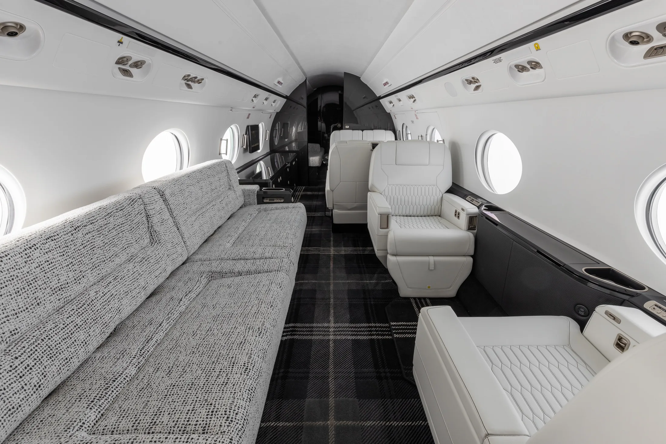 The elegant interior cabin of a private jet, showing a long grey sofa, white seats, and a dark plaid carpet.