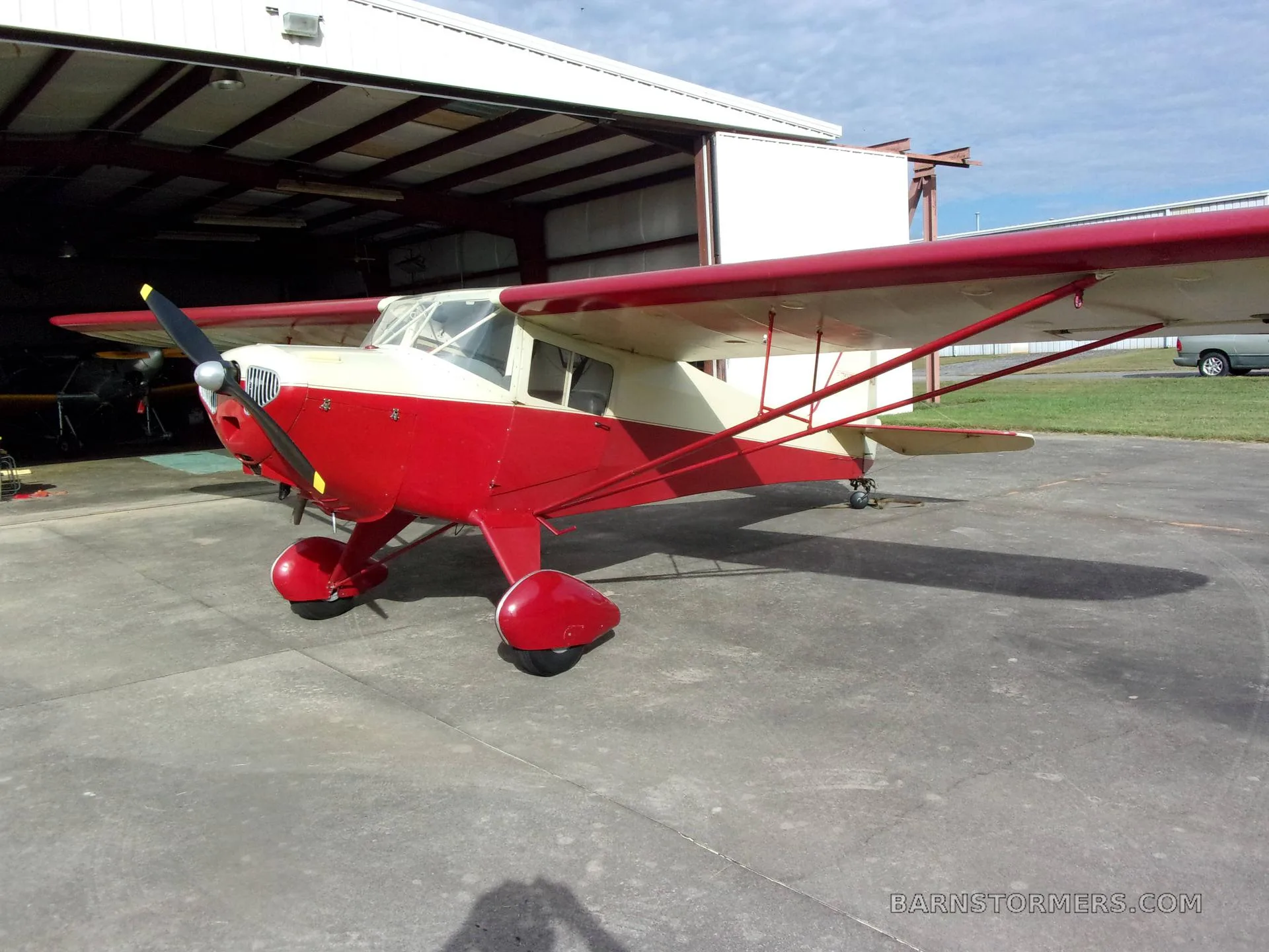 A red and cream Taylorcraft BC-12D aircraft parked on a concrete apron outside a hangar, viewed from the front-left.