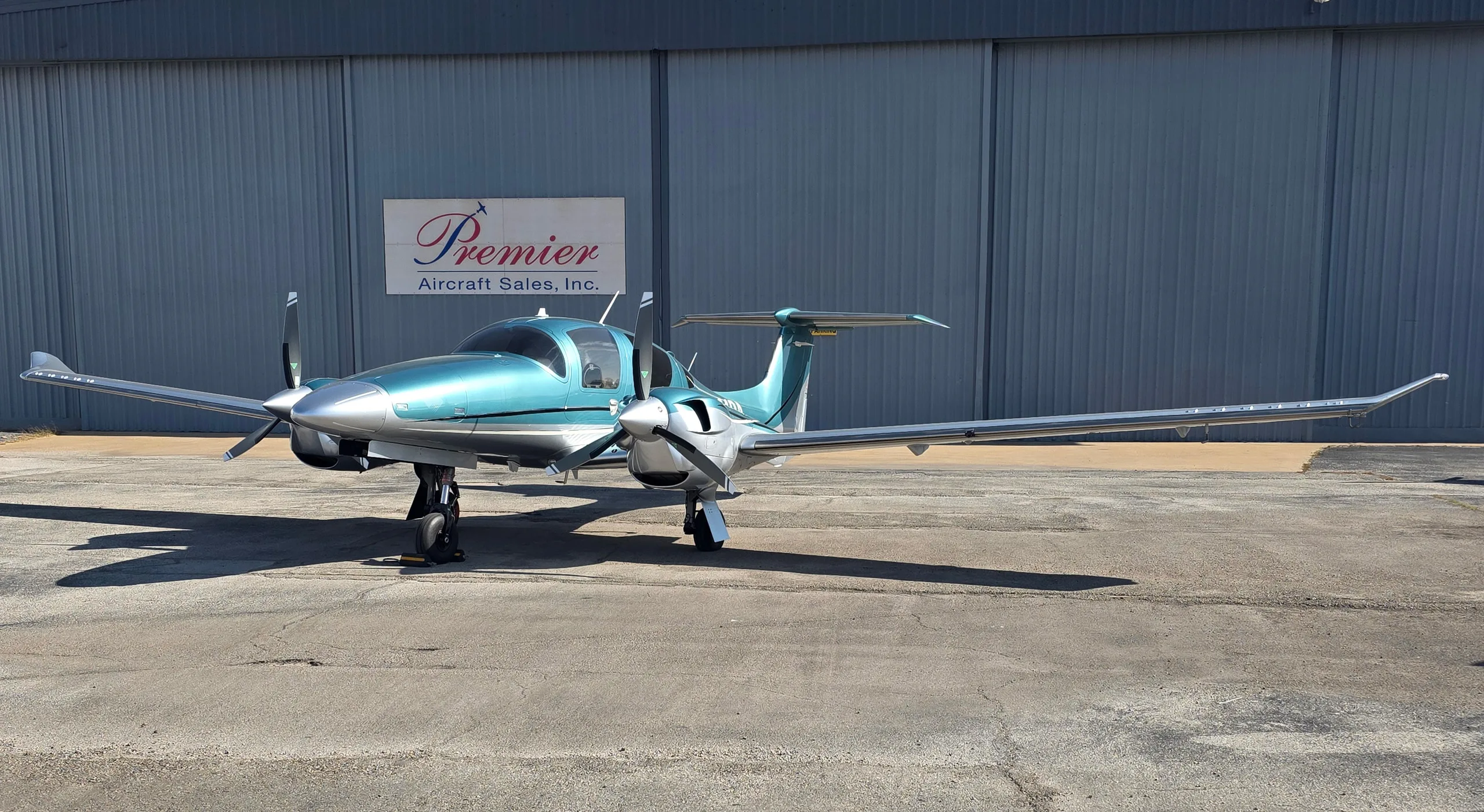 A teal and silver Diamond DA62 twin-engine aircraft parked on an asphalt ramp in front of a hangar.