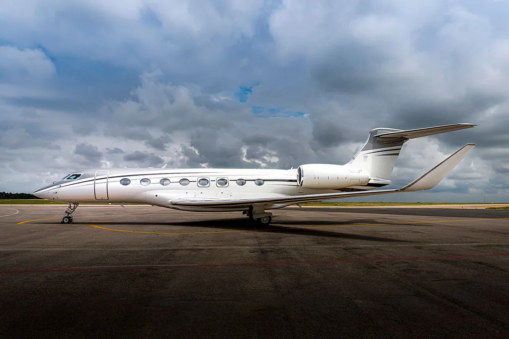 A white Gulfstream G650 business jet with black accent stripes parked on an airport tarmac under a cloudy sky.