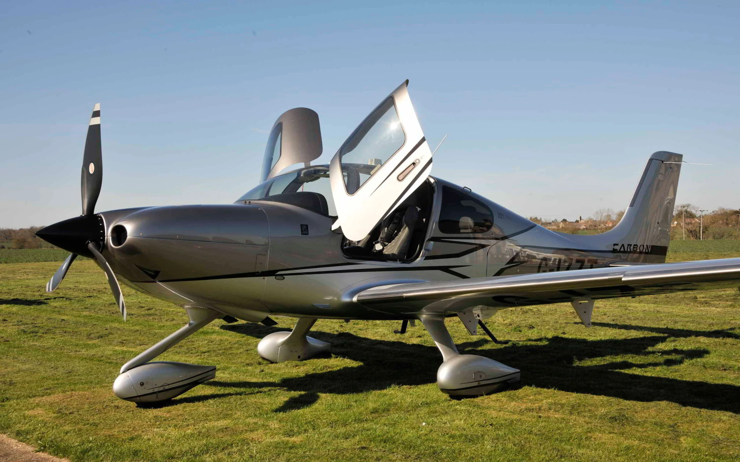 A silver Cirrus SR22T aircraft with open doors parked on a grassy field under a clear blue sky.