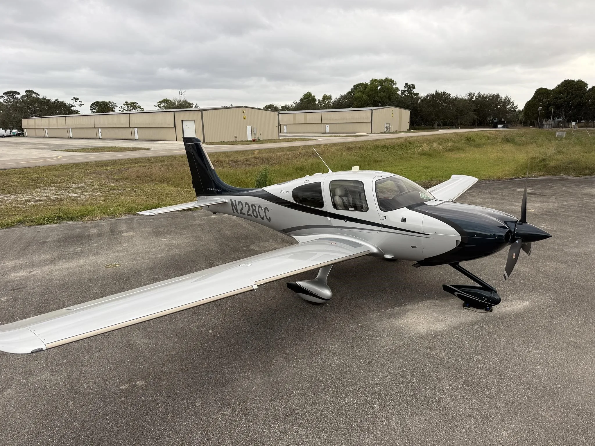 A Cirrus SR22T aircraft, N228CC, with a black and silver paint scheme, parked on an asphalt ramp under a cloudy sky.