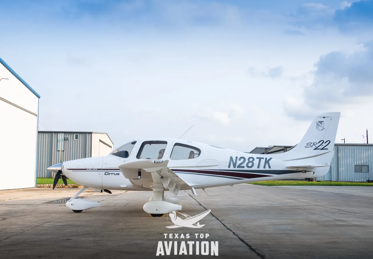 A side view of a white Cirrus SR22 aircraft with maroon and gold stripes, registration N28TK, parked on a tarmac next to a hangar.
