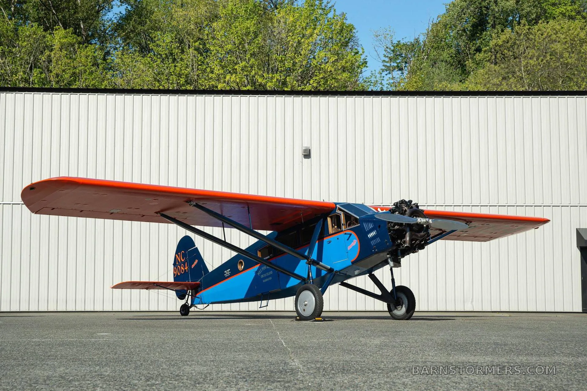 A vintage blue and orange high-wing monoplane, registration NC 9084, parked on an airfield in front of a white hangar.