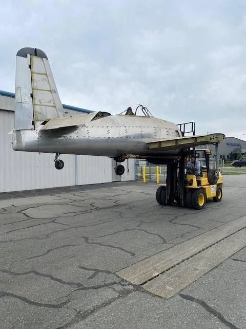 A bare metal aircraft fuselage, missing wings and engine, is being moved by a yellow forklift on an asphalt tarmac.
