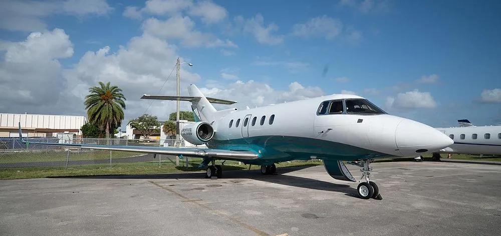 A Hawker 800XP business jet with a white and teal paint scheme parked on an airfield under a blue sky.