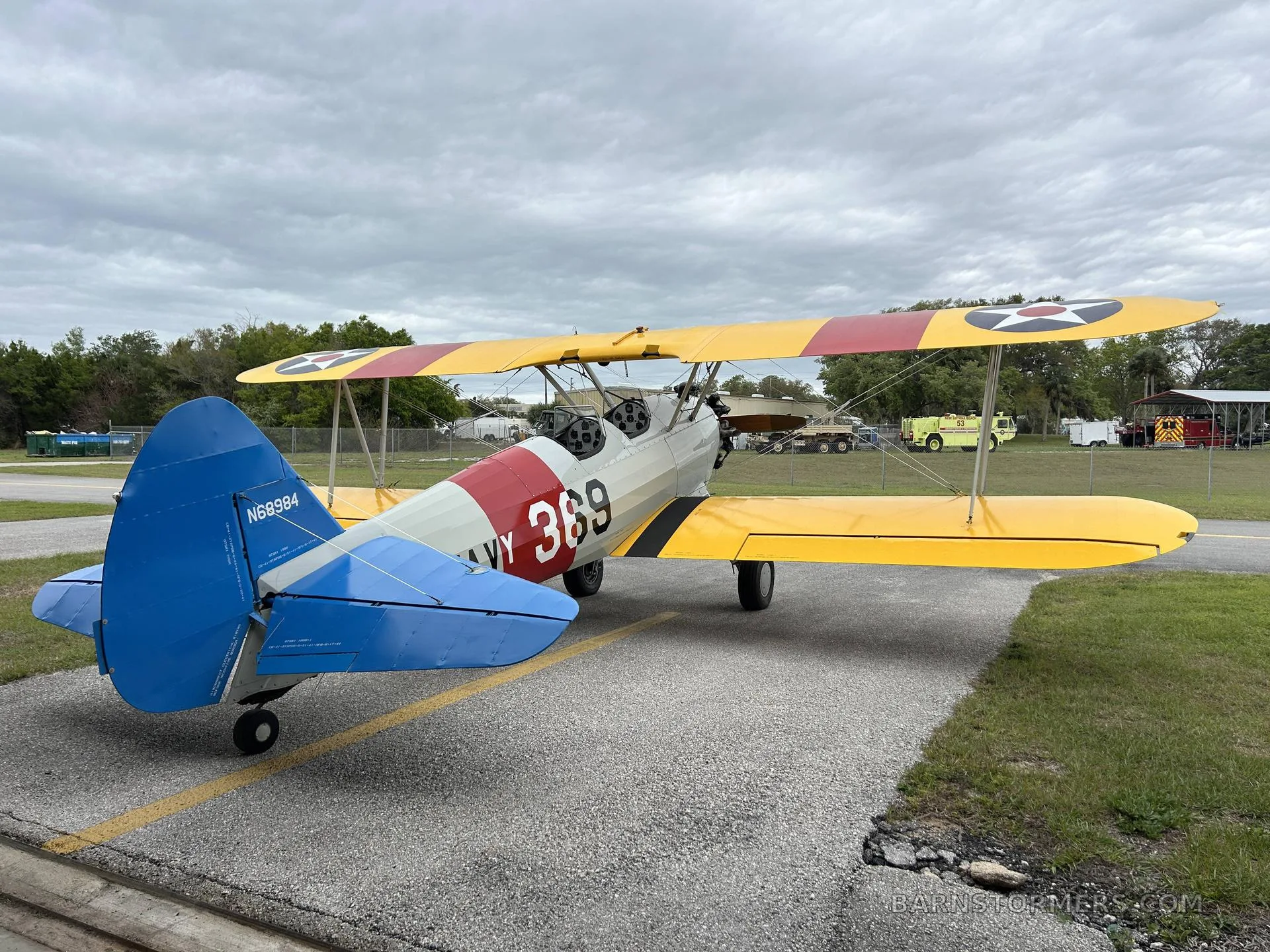 A yellow and white biplane with 'US NAVY 369' markings and a blue tail, seen from the rear-left on an asphalt ramp.