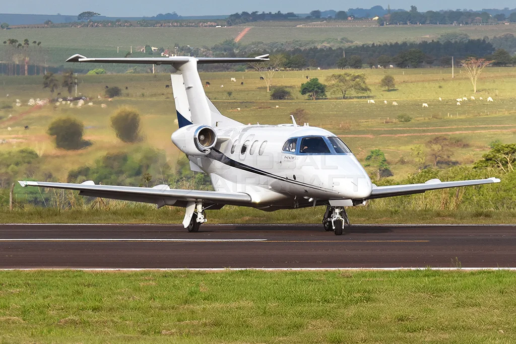 A white Embraer Phenom 100 with blue and black stripes on a runway, with a grassy, hilly background.